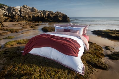 Celso de Lemos BAYA pillowcase: A bed with a red throw and striped pillowcases on a rocky seashore