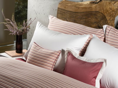 Celso de Lemos BAYA pillowcase: White and striped pillows and a pink cushion on a bed, with a vase of dried flowers behind