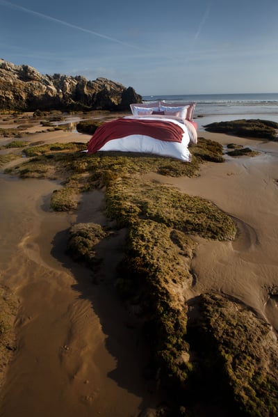 Celso de Lemos BAYA pillowcase: A bed with a red throw on a beach, with rocks and the sea behind
