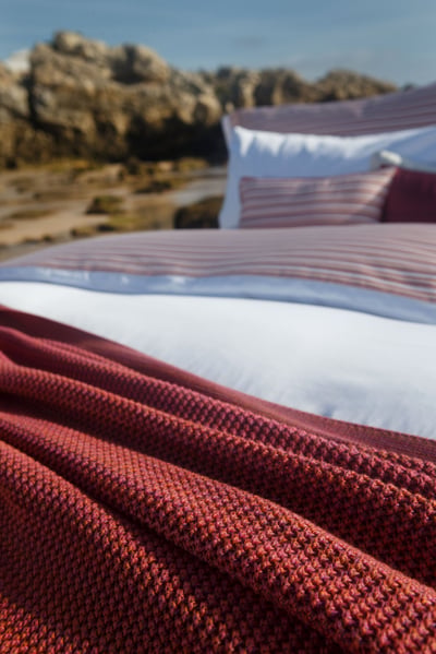 Celso de Lemos BAYA pillowcase: The foot corner of a bed with a red knitted throw on a beach, striped pillows behind