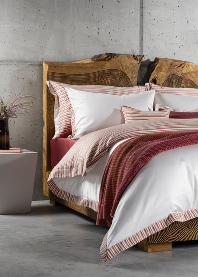 Celso de Lemos BAYA pillowcase: A bed in front of a concrete wall and a wooden headboard, dressed in white and striped linen with a red throw