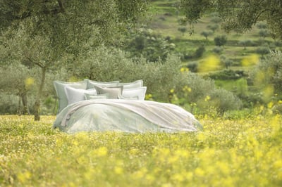 Celso de Lemos BIJOUX pillowcase: A bed in pale green and white linen under olive trees on a flowering meadow