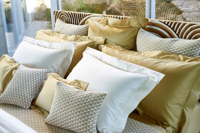 Celso de Lemos BOURDON duvet cover: A pile of gold and white pillows and patterned cushions in front of a glass wall